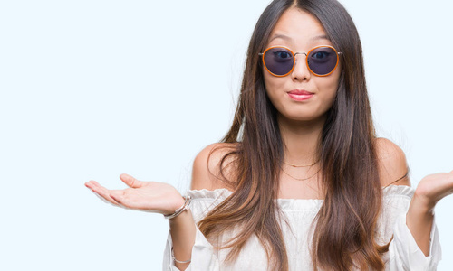 年轻的亚洲女人戴着太阳镜，在孤立的背景上毫无头绪，表情混乱，举起手臂和手。 怀疑概念。