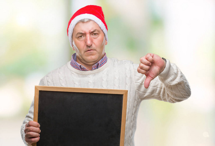 英俊的老人戴着圣诞帽，在孤立的背景上拿着黑板，脸上带着愤怒的消极迹象，表示不喜欢，竖起大拇指拒绝概念
