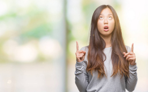 年轻的亚洲女人在孤立的背景下惊讶和惊讶地抬头，用手指和举起的手臂指着。