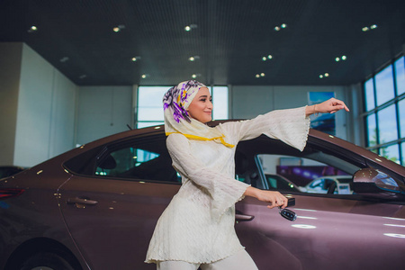 财产和人概念穆斯林妇女在头巾与汽车钥匙在汽车显示背景。快乐的女人从经销商在车展或沙龙的汽车钥匙