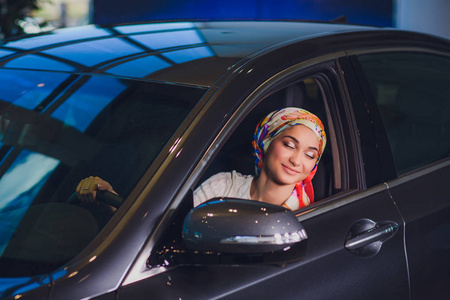 财产和人概念穆斯林妇女在头巾与汽车钥匙在汽车显示背景。快乐的女人从经销商在车展或沙龙的汽车钥匙