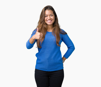 年轻漂亮的黑发女人穿着蓝色毛衣，在孤立的背景下，用手做快乐的竖起大拇指的手势。 赞许的表情看着相机显示成功。