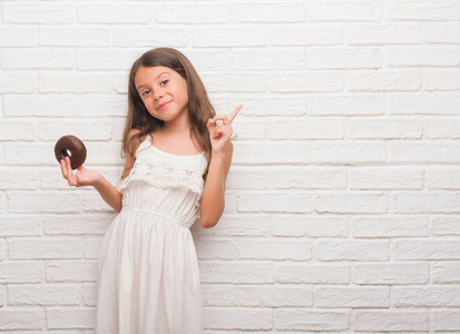 年轻的西班牙裔小孩在白色砖墙上吃巧克力甜甜圈，非常高兴地用手和手指指向一边