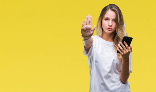年轻漂亮的金发女人使用智能手机在孤立的背景下，张开手做停止标志，严肃自信的表情防御手势