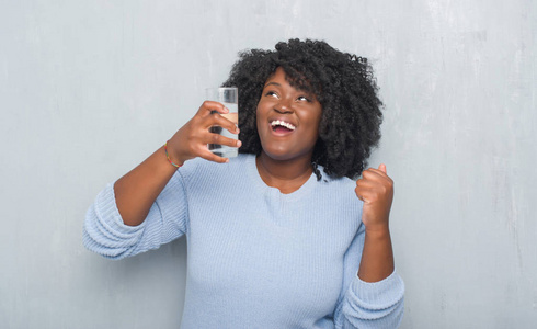 年轻的非洲裔美国妇女在灰色的格朗格墙上喝着一杯水，尖叫自豪，庆祝胜利和成功，非常兴奋，欢呼的情绪