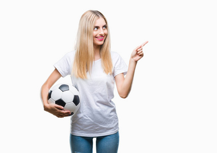 年轻漂亮的金发女郎把足球抱在孤立的背景上，非常高兴地用手和手指指向一边