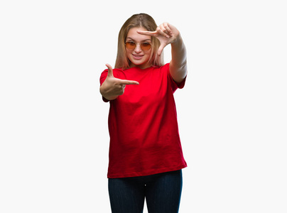 年轻的白种人女人戴着太阳镜，在孤立的背景上微笑，用手和手指制作框架，用快乐的脸。 创造力和摄影理念。