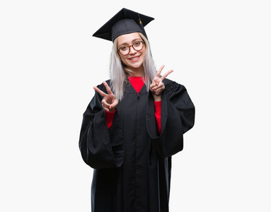 年轻的金发女人穿着研究生制服，在孤立的背景上微笑着，看着镜头，手指在做胜利的标志。 二号。