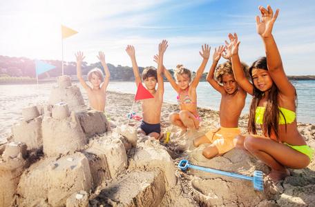 一大群多民族的孩子，快乐的朋友坐在沙滩上，围着沙堡挥舞着手
