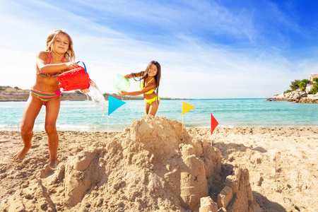 两个穿比基尼的快乐女孩的肖像，从桶里倒水，在海滩上建造沙堡