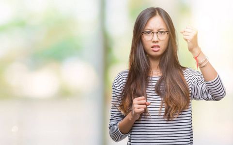 年轻的亚洲女人戴着眼镜，在孤立的背景下，愤怒和疯狂地举起拳头，沮丧和愤怒，同时大声喊叫。 愤怒和侵略性的概念。