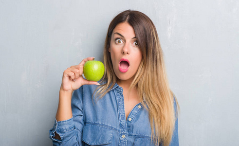 一个年轻的成年女人在灰色的墙上吃着新鲜的绿色苹果，吓得满脸惊讶，害怕，兴奋的表情