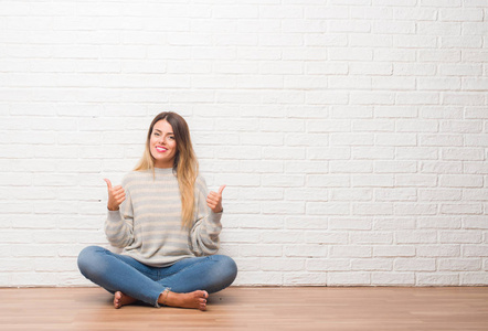 年轻的成年女人坐在地板上，在白色砖墙上，在家里做积极的手势，用手竖起大拇指，微笑和快乐。 用欢快的表情看着相机，胜利者的手势。