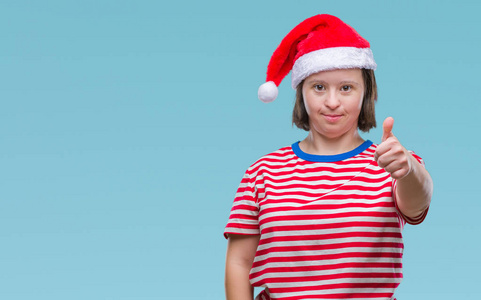 年轻的成年妇女患有唐氏综合症，戴着圣诞帽，在孤立的背景下，用手做快乐的拇指向上的手势。 赞许的表情看着相机，显示出成功。