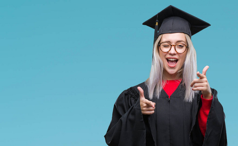 年轻的金发女人穿着研究生制服，在孤立的背景上指着相机，带着快乐和有趣的脸。 良好的能量和氛围。