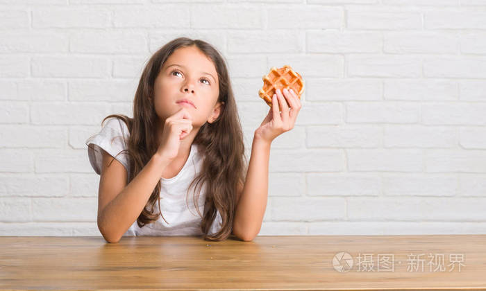 年轻的西班牙裔小孩坐在桌子上吃华夫饼严肃的脸思考