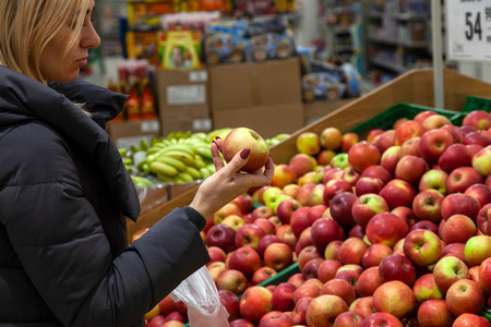 一个年轻的金发女孩穿着一件长夹克，在水果部门的一家商场购物时选择食物，手里拿着一个红绿色的苹果，手里拿着许多成熟的产品。