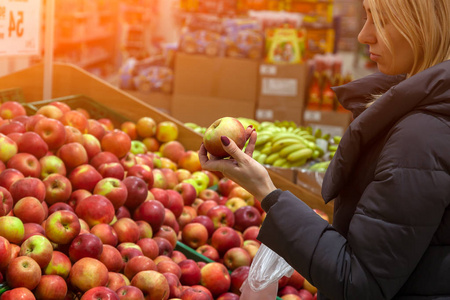 一个年轻的金发美女穿着一件长夹克，在水果部门的一家商场购物时选择食物，手里拿着一个红绿色的苹果，手里拿着许多成熟的产品。