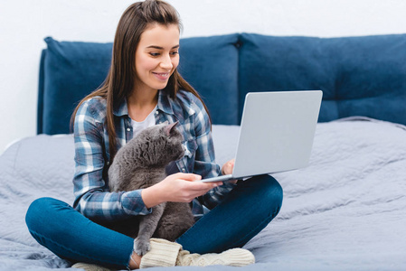 带着可爱的英国短毛猫坐在床上，用笔记本电脑微笑的年轻女人