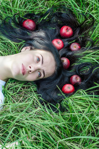 迷人的女孩，黑发躺在草地上的绿色草地上。 地上的头发。 头发上的红苹果。 夏天的心情回忆。 自然营养健康。