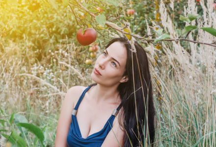 一个穿着夏装的女人坐在苹果园的枯草里，看着树枝上的一个红苹果。 引诱。 妇女健康。 生殖。 孩子的诞生。