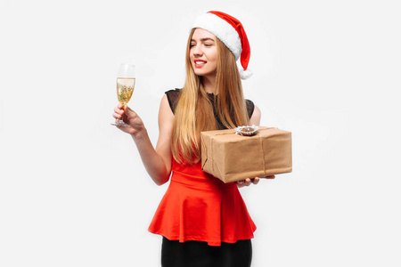 快乐，年轻的女人穿着连衣裙和圣诞老人的帽子，庆祝新年，在她的手中，一杯香槟和一份圣诞礼物。在白色背景上。圣诞节的概念。