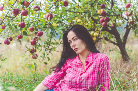 穿着格子粉色衬衫的年轻女人坐在一棵大红苹果树下的一个老苹果园里。收获。健康食品。秋天的心情。