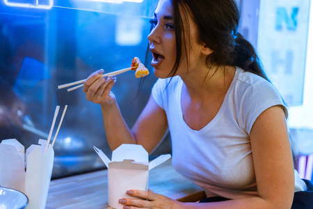 在咖啡馆或餐馆用筷子吃亚洲食物的年轻有魅力的女人。女人用传统的白菜棒吃米饭和炸鸡。