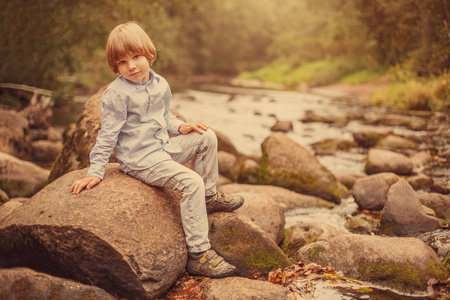 一个男孩在自然背景上的肖像。 那孩子坐在河边的岩石上
