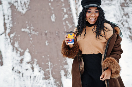 穿着羊皮外套和帽子的非裔美国妇女在冬日的雪地背景下，用一杯咖啡摆好姿势。