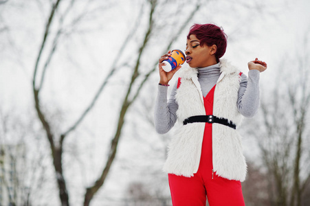 穿着红色裤子和白色毛皮外套的非洲裔美国妇女在冬日的雪地背景下摆姿势，拿着一杯咖啡。