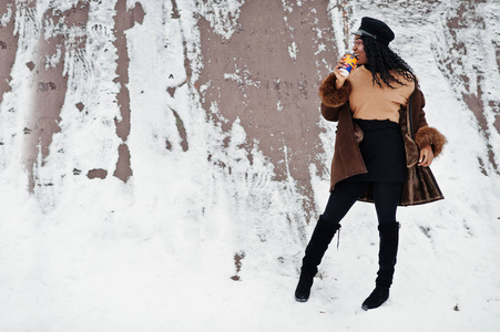 穿着羊皮外套和帽子的非裔美国妇女在冬日的雪地背景下拿着一杯咖啡摆姿势。