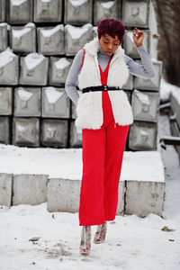 穿着红色裤子和白色毛皮外套的非洲裔美国妇女在冬日的雪地石背景下摆好姿势。