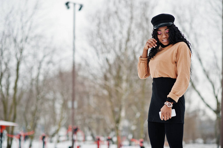 穿着黑色裙子棕色毛衣和帽子的非裔美国妇女在冬日的雪地背景下摆好姿势，手里拿着电话。