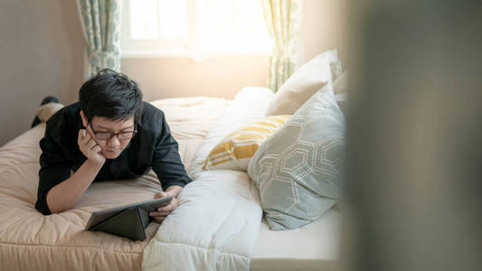 年轻的亚洲商人在舒适的卧室里躺在床上时使用数字平板电脑和钢笔。 家庭生活方式与现代电子产品概念