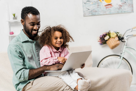 非裔美国人父亲和可爱的孩子坐在沙发上，用笔记本电脑