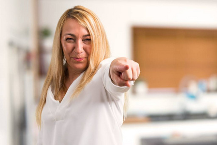 中年金发女人穿着白色衬衫，愤怒地愤怒着。 对糟糕的情况感到沮丧，并指着家里的前面