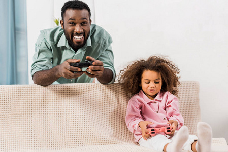 非裔美国人爸爸站着和女儿玩电子游戏，客厅里