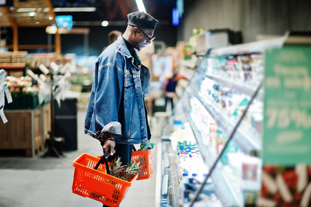 时尚休闲的非裔美国人男人穿着牛仔裤夹克和黑色贝雷帽，拿着两个篮子站在冰箱附近，在超市购物。