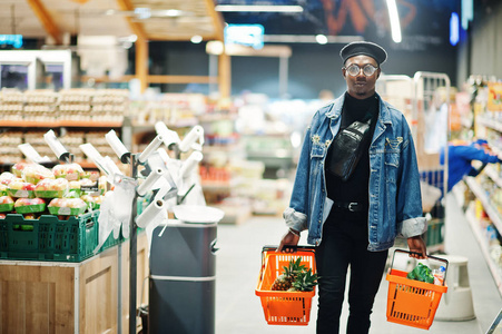 时尚休闲的非裔美国人男人穿着牛仔裤夹克和黑色贝雷帽，拿着两个篮子在超市散步和购物。
