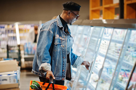时尚休闲的非裔美国人男子，穿着牛仔裤夹克和黑色贝雷帽，拿着篮子站在美食冰箱附近，在超市购物。