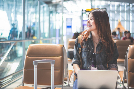 美丽的亚洲女人使用笔记本电脑，看着外面的机场。 坐在机场航站楼的女人。 人和生活方式的概念。 技术和旅游主题。 商业和肖像主题。