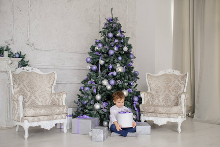 一个6岁的金发男孩，手里拿着礼物，靠近一棵圣诞树，上面有紫色的玩具和两把白色的老式扶手椅