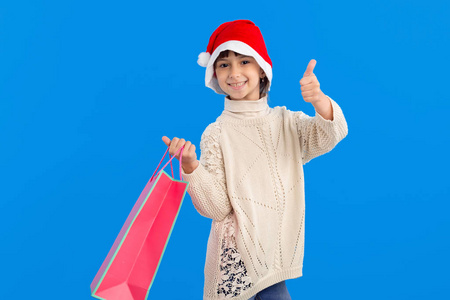 就像漂亮的女孩穿着冬季夹克和圣诞老人帽子，蓝色背景上有一个粉红色的礼品袋。竖起大拇指圣诞节和新年的好购物。