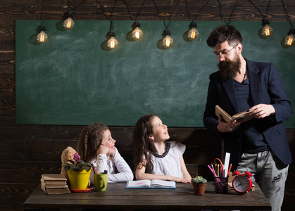老师和女孩学生在教室, 黑板上的背景。留胡子的男人教女学生看书。好奇开朗的孩子听老师的注意。小学理念