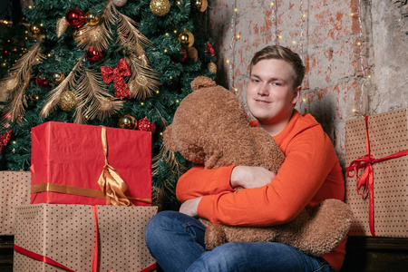 帅哥抱着熊坐在树下，周围是礼物