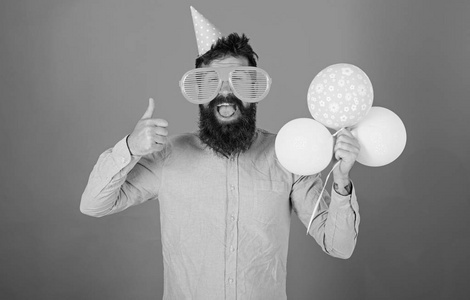 时髦的大太阳镜庆祝生日。脸上留着胡子和胡须的男人抱着一堆气球, 红色的背景。庆祝概念。人在党帽子显示拇指向上手势