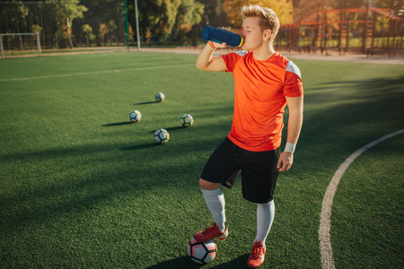 年轻足球运动员站在草坪上喝水的照片。他把脚放在球上。后面还有四个球乌斯蒂德阳光明媚的夏季天气