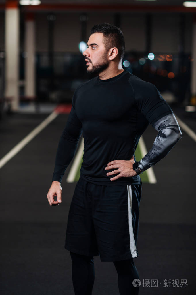 英俊的男人,肌肉发达,穿着黑色衣服在体育馆的镜头前摆姿势