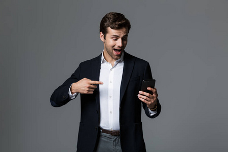 一位年轻的快乐商人的肖像，他穿着西装，被孤立在灰色背景上，手指着手机